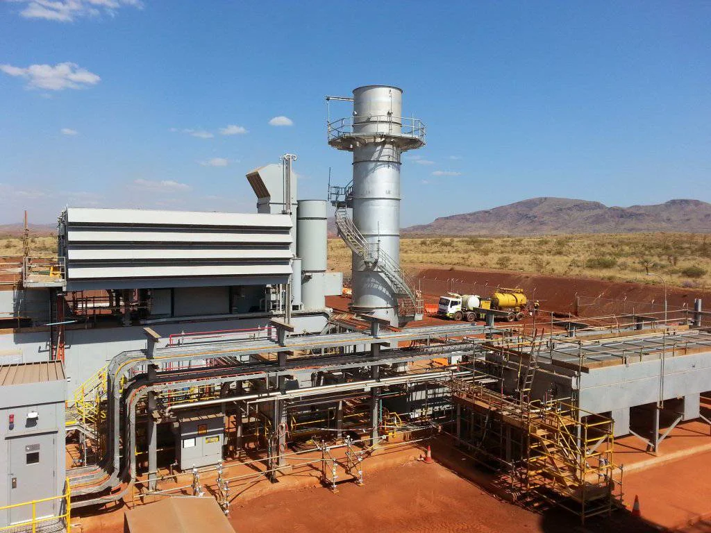 A power plant facility with various pipes, metal structures, and a tall exhaust stack, set in a dry, open landscape with mountains in the distance under a clear blue sky.